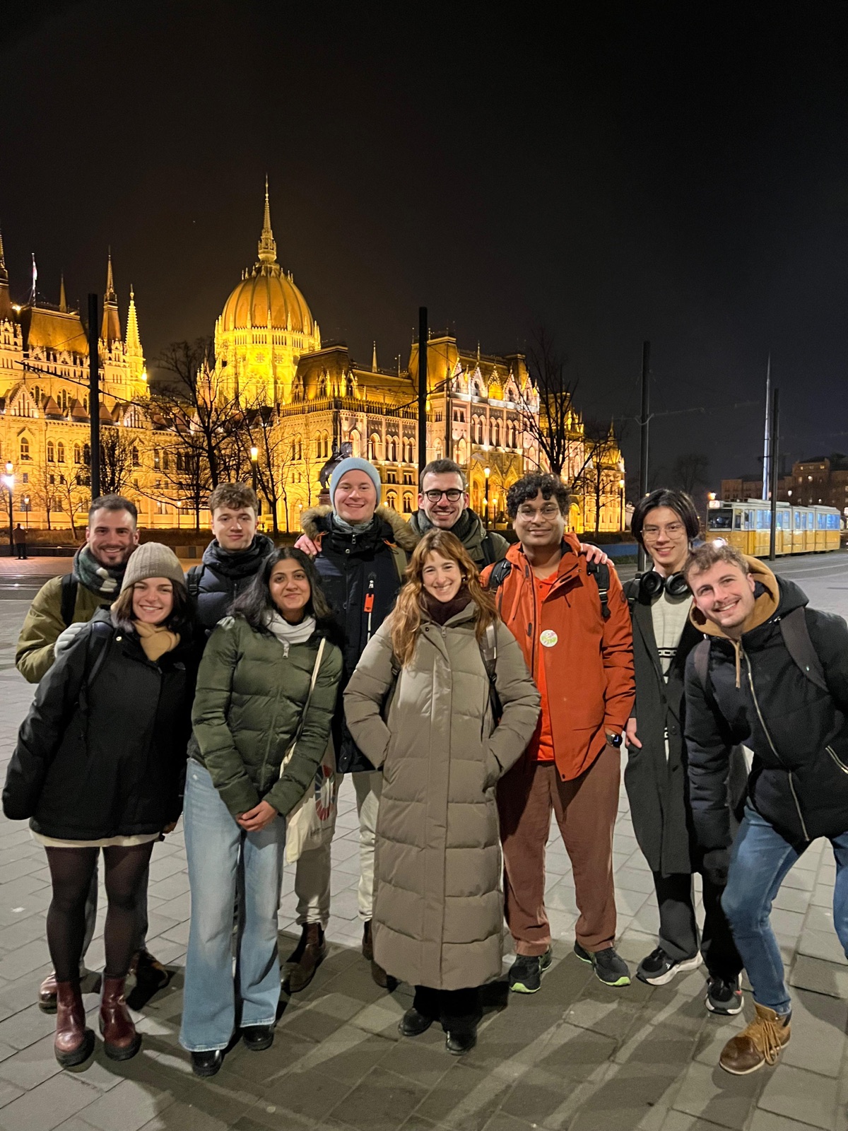 Ten people in winter coats in front of the lit Hungarian Parliament Building at night